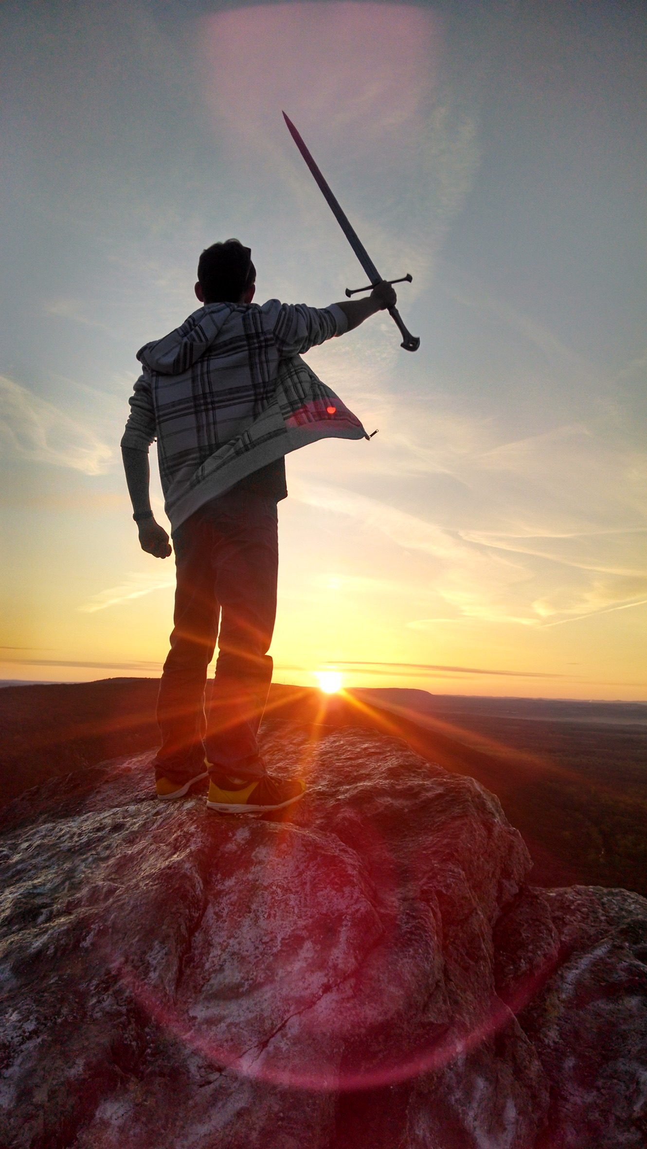 Dalton on a mountain summit at sunset with a sword raised toward the sky